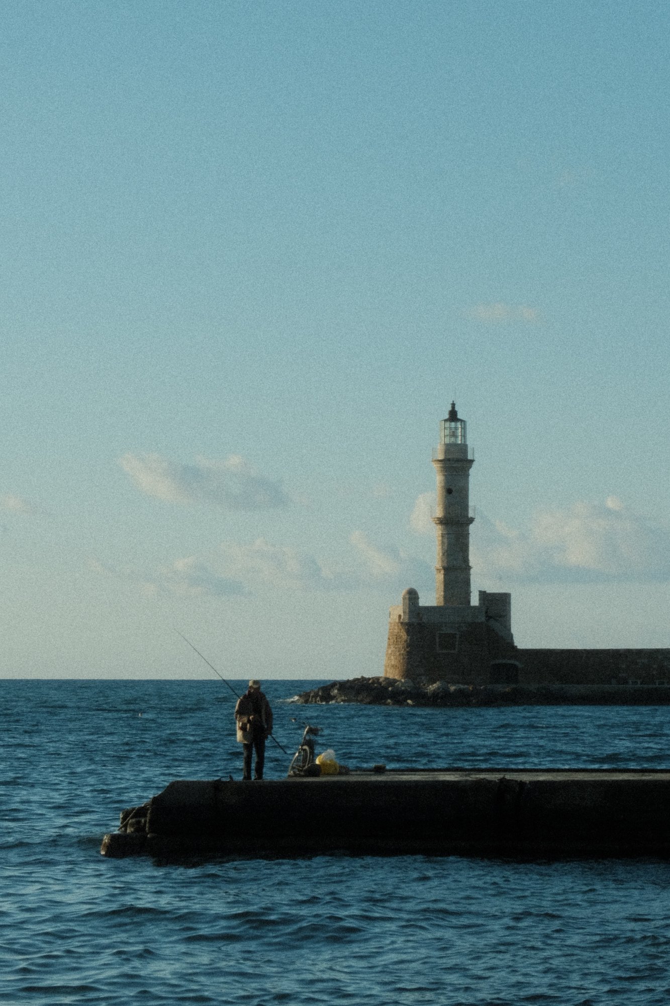 A fisherman standing on a jetty with the lighthouse of Chania, Greece in the background