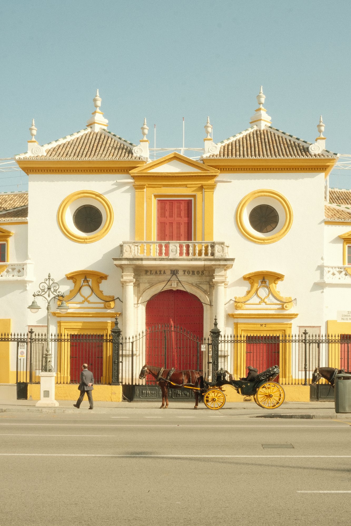 The ornate white and yellow facade of the Plaza de Toros bullring in Seville, Spain, with a horse-drawn carriage out front